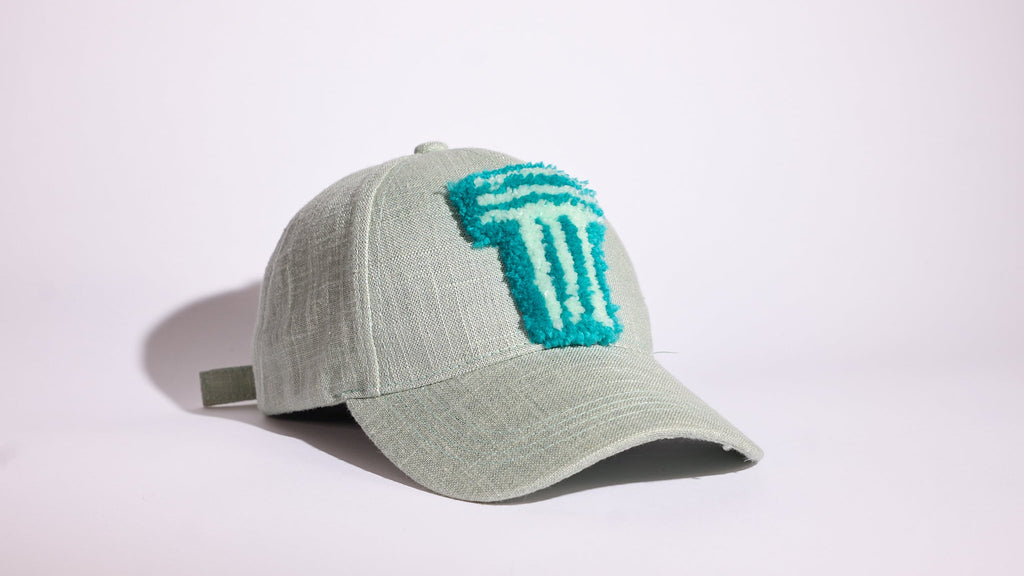 A light sage green baseball cap featuring a textured turquoise embroidered logo on the front, set against a plain white studio background.