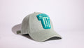 A light sage green baseball cap featuring a textured turquoise embroidered logo on the front, set against a plain white studio background.