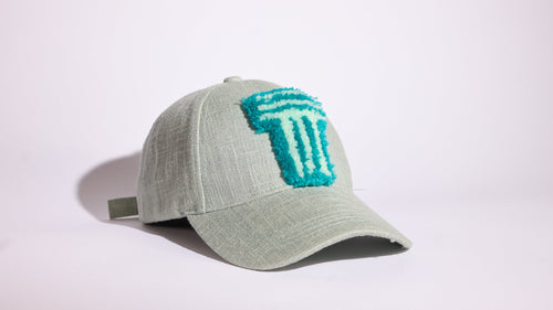 A light sage green baseball cap featuring a textured turquoise embroidered logo on the front, set against a plain white studio background.