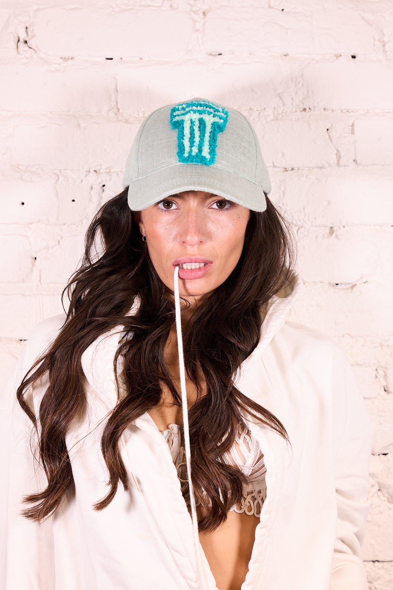 A woman with long wavy brown hair wearing a light gray baseball cap with a turquoise embroidered 'T' logo, posing against a white brick wall.
