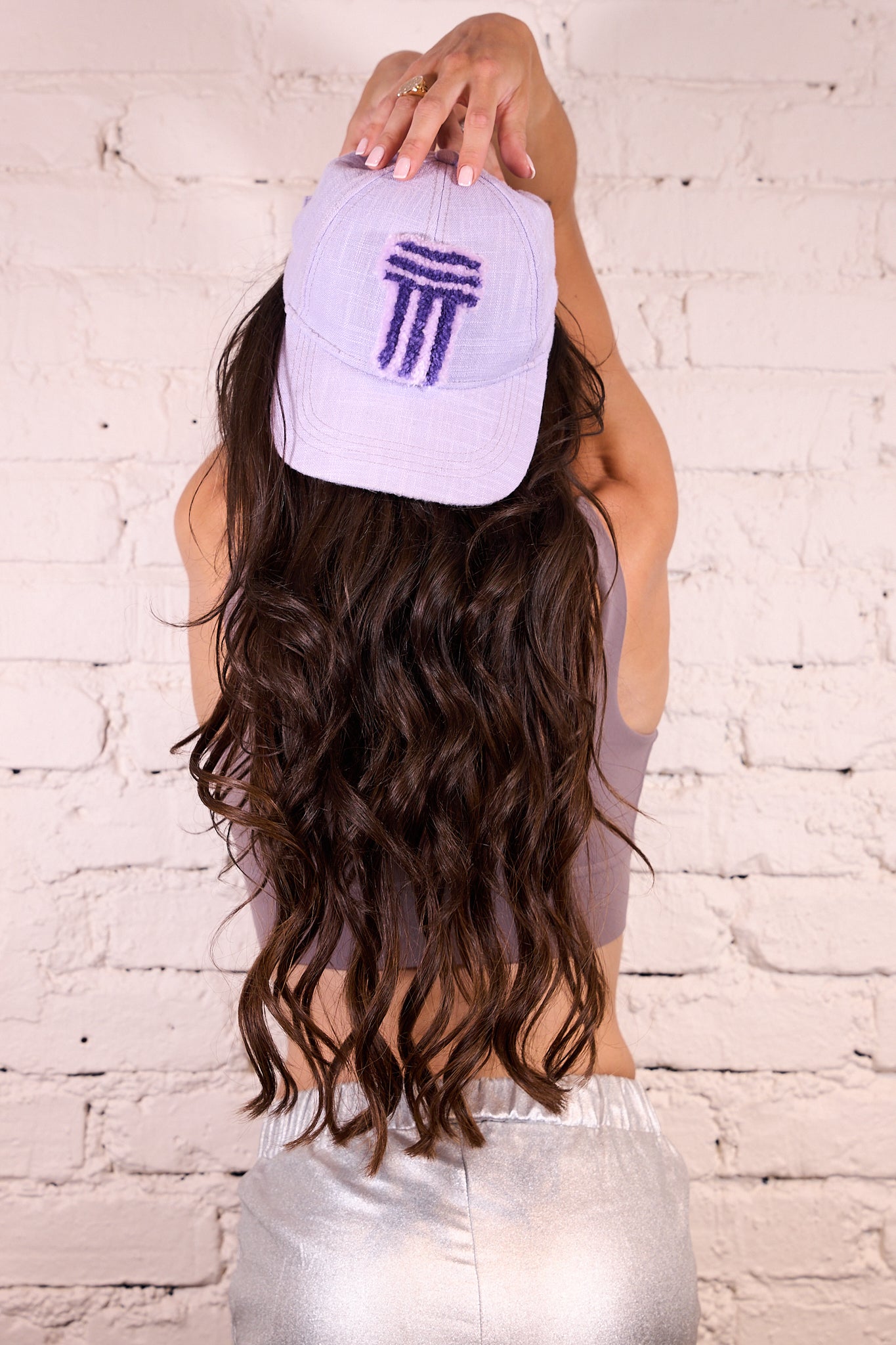 A woman with long wavy brown hair seen from behind, wearing a light lavender baseball cap with a dark purple embroidered logo, a mauve crop top, and shiny silver pants, standing against a white brick wall.