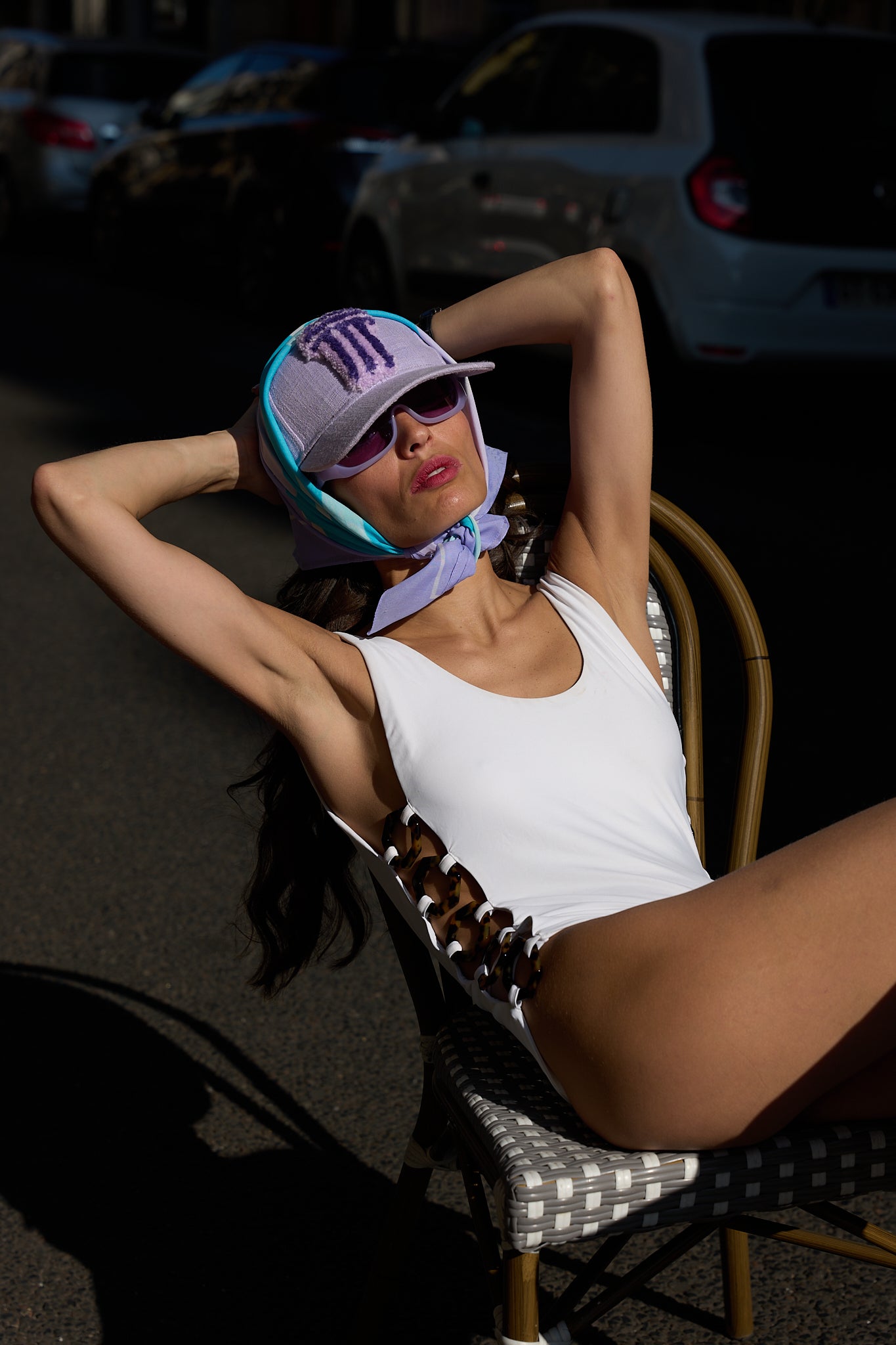 A woman reclining on a chair outdoors, wearing a white bodysuit, a light purple Tufted  baseball cap, and sunglasses. The background shows parked cars in a street setting.