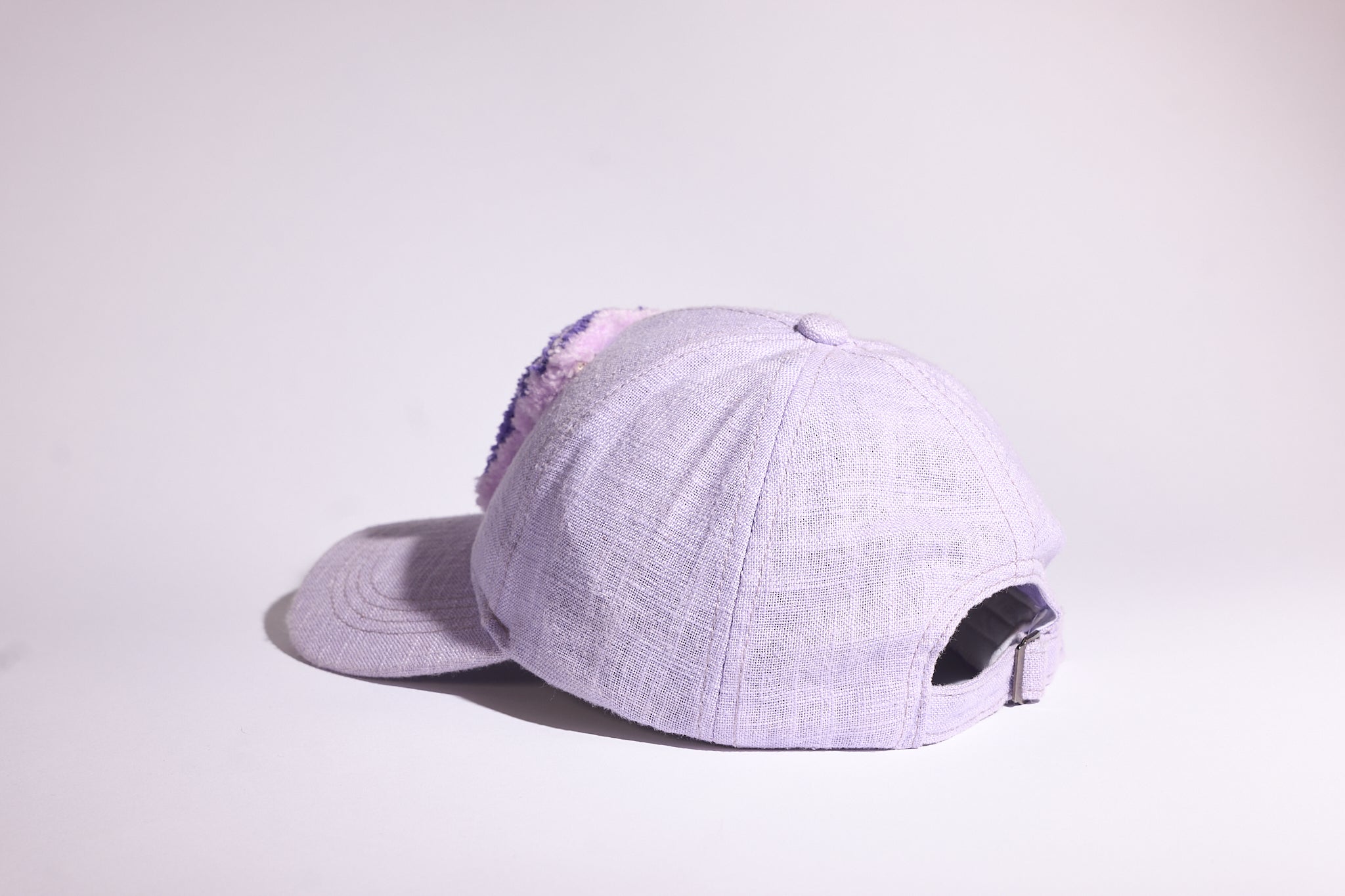 A light lavender baseball cap with a textured linen-like fabric, featuring a purple sequined embellishment on the side and a black metallic adjustment buckle on the back, set against a plain light gray background.