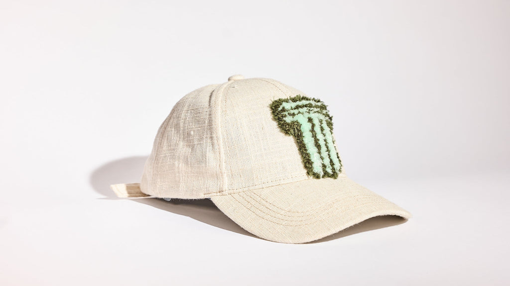 A light beige linen baseball cap featuring a green beaded 'T' patch on the front, photographed on a clean white studio background.