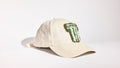 A light beige linen baseball cap featuring a green beaded 'T' patch on the front, photographed on a clean white studio background.