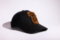 A black baseball cap featuring a textured, fuzzy brown and black embroidered tuft on the front, photographed on a clean white background with a soft shadow.