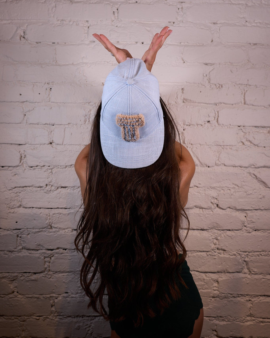 A woman with long dark hair poses from behind against a white brick wall, wearing a light blue baseball cap with a graphic patch on the back and a black swimsuit.