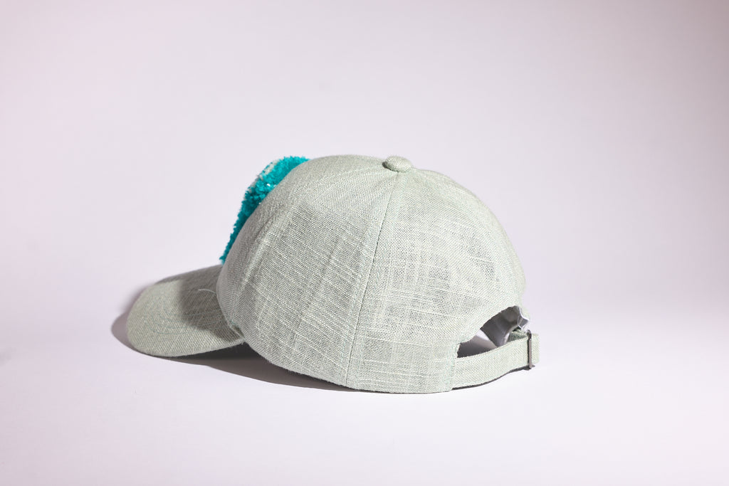 A light sage green textured baseball cap with a bright turquoise blue fuzzy trim on the back, shown from a side profile against a clean white studio background.