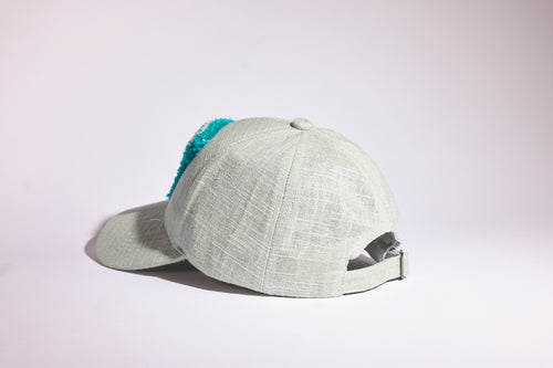 A light sage green textured baseball cap with a bright turquoise blue fuzzy trim on the back, shown from a side profile against a clean white studio background.