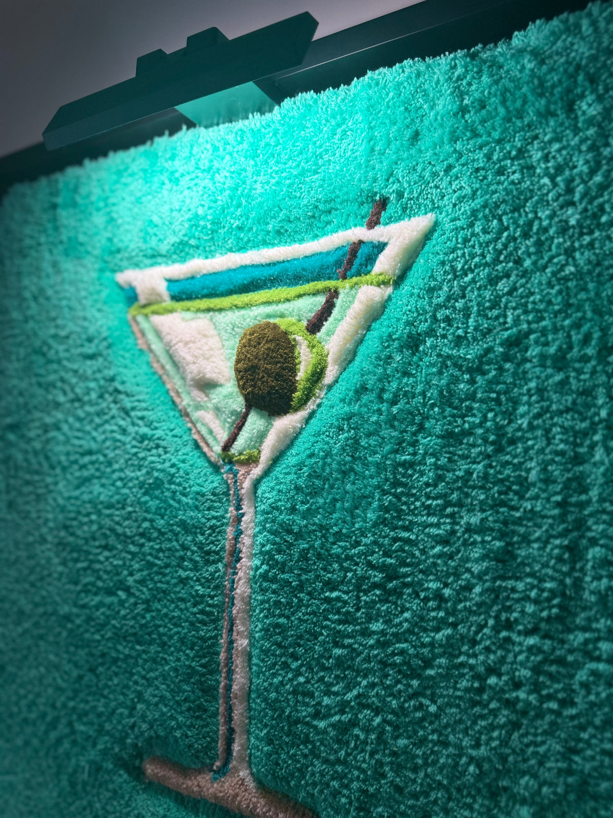 VESPER, œuvre tuftée contemporaine de Ninon Dechavanne, vue en perspective sous éclairage pour révéler le relief du verre à cocktail et de l’olive.